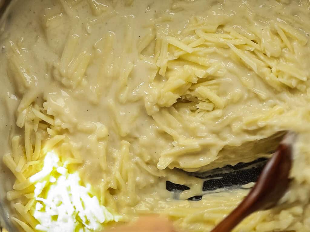Cheesy Mushroom Rigatoni Pie - Easy Vegan Recipe. 34 Close-up of shredded cheese being stirred into a creamy white sauce with a wooden spoon.