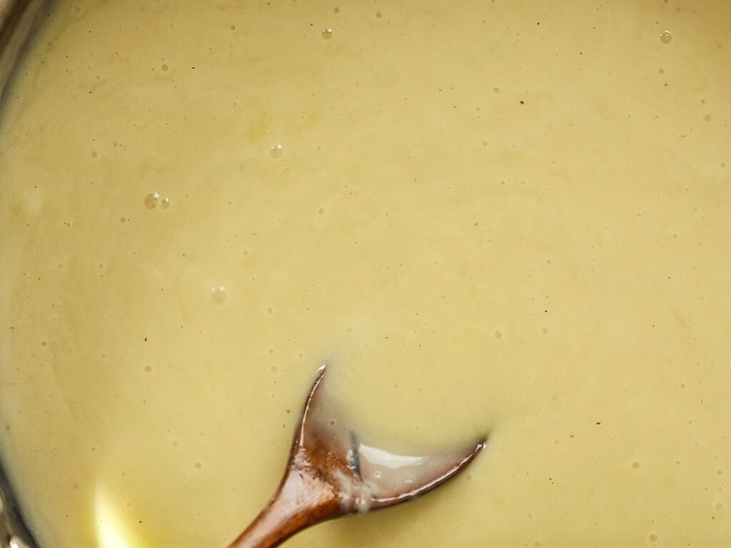 Cheesy Mushroom Rigatoni Pie - Easy Vegan Recipe. 35 A close-up of a creamy, light yellow sauce being stirred with a wooden spoon. The texture appears smooth with a few small bubbles on the surface.