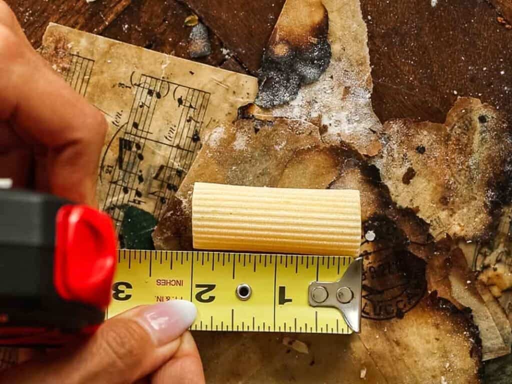 Cheesy Mushroom Rigatoni Pie - Easy Vegan Recipe. 5 A hand holds a tape measure against a small wooden dowel on a cluttered, stained surface with old, burnt paper and sheet music in the background.