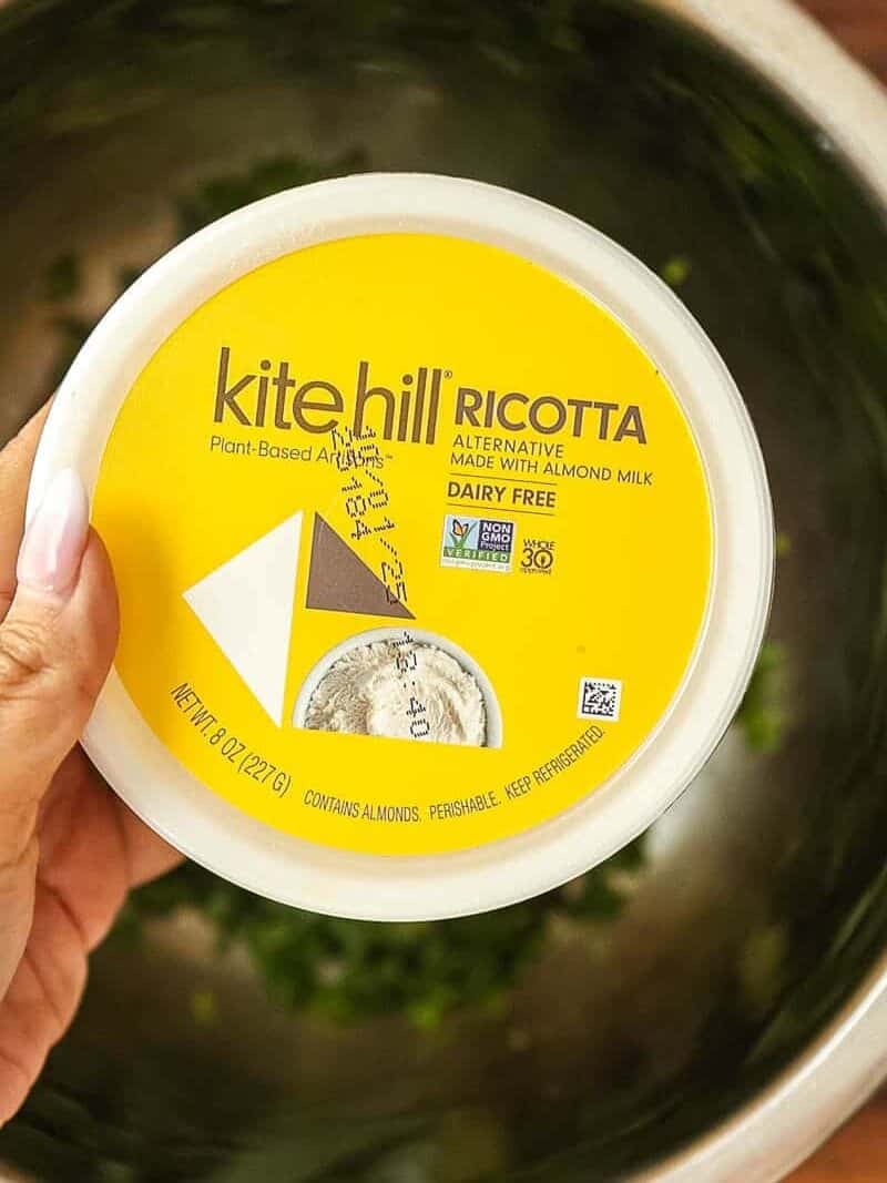 Cheesy Mushroom Rigatoni Pie - Easy Vegan Recipe. 12 A hand holds a container of Kite Hill Ricotta, a plant-based, dairy-free cheese made from almond milk, over a metal bowl with chopped green herbs inside.