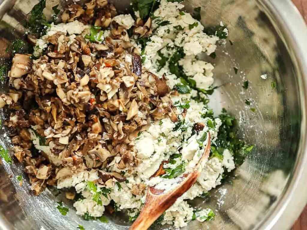 Cheesy Mushroom Rigatoni Pie - Easy Vegan Recipe. 37 A metal bowl contains a mixture of chopped mushrooms, ricotta, and fresh herbs being mixed with a wooden spoon.
