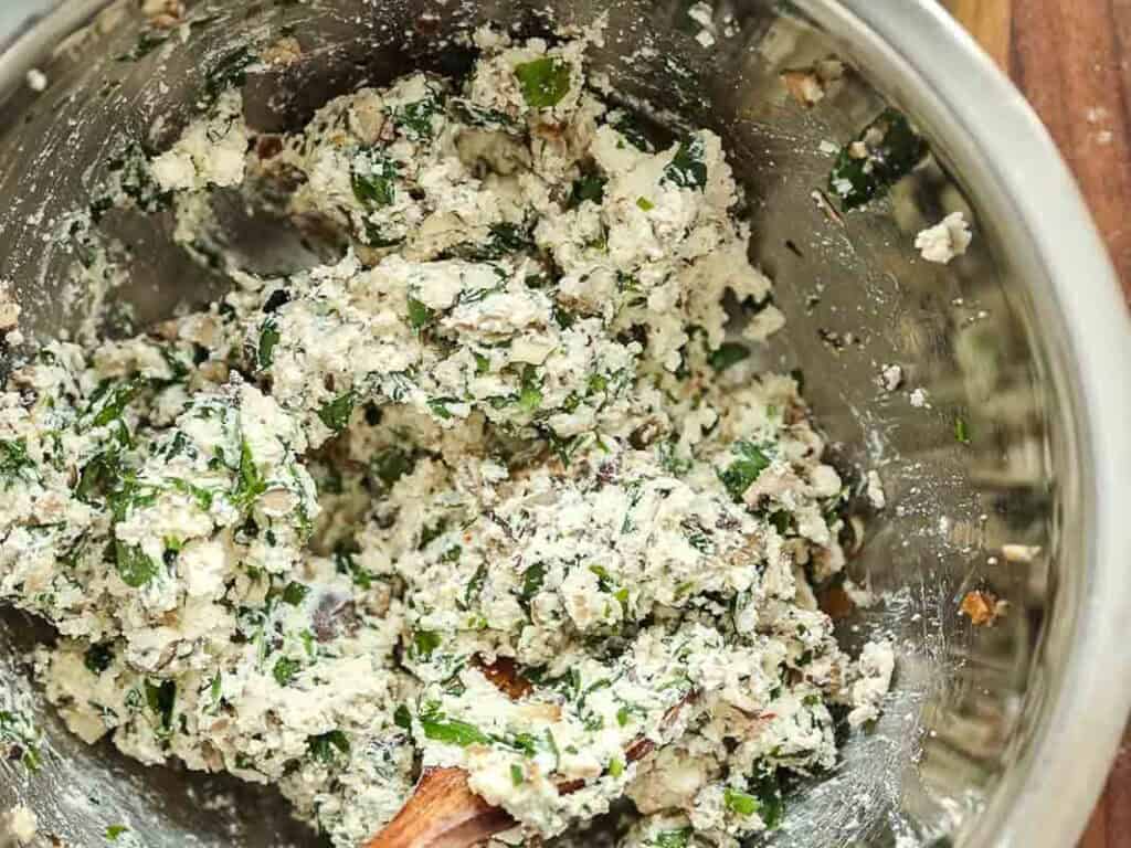 Cheesy Mushroom Rigatoni Pie - Easy Vegan Recipe. 39 A metal mixing bowl containing a mixture of crumbled cheese, chopped greens, and herbs being combined with a wooden spoon.