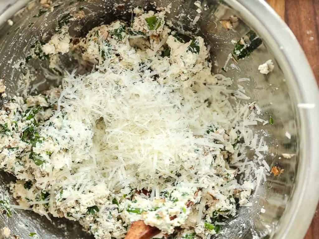 Cheesy Mushroom Rigatoni Pie - Easy Vegan Recipe. 38 A metal mixing bowl filled with a mixture of ricotta cheese, herbs, and shredded parmesan cheese. A wooden spoon is partially visible in the bowl.