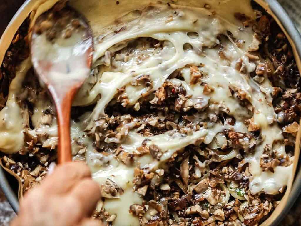 Cheesy Mushroom Rigatoni Pie - Easy Vegan Recipe. 48 a hand holding wooden spoon mixes béchamel with minced mushroom mixture.