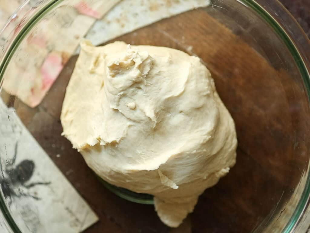 How to Make Viral Salt Bread Recipe (Easy and Vegan). 13 A ball of pale, smooth bread dough sits in a clear glass bowl on a wooden surface with paper scraps nearby.