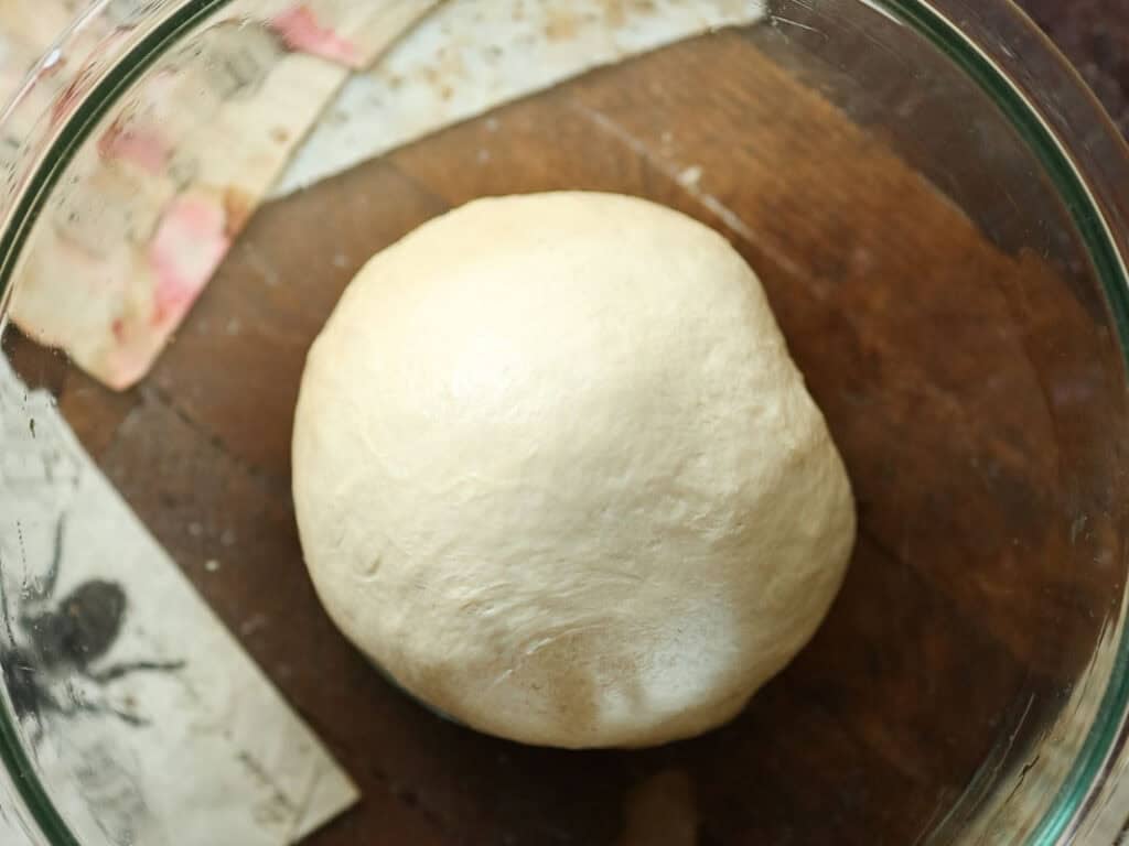 How to Make Viral Salt Bread Recipe (Easy and Vegan). 14 A smooth ball of dough rests in a clear glass bowl on a wooden surface, with pieces of paper partially visible beneath the bowl.