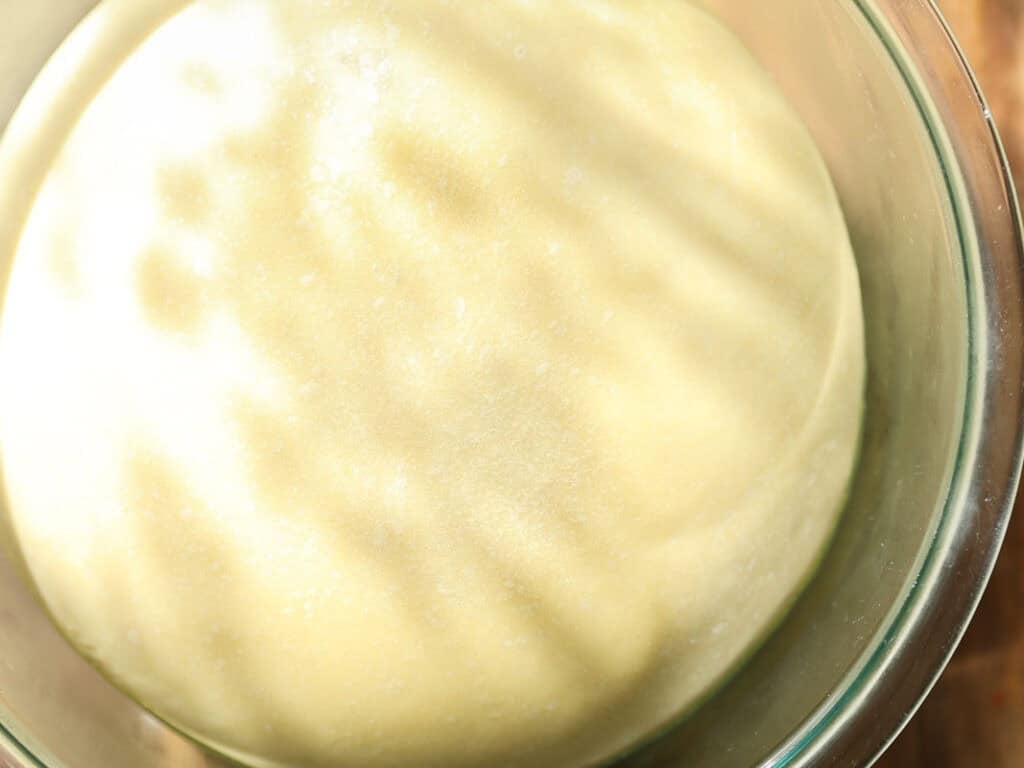 Easy and Delicious Garlic Knots Recipe (Vegan) 10 A close-up of risen dough in a glass bowl, with sunlight creating soft shadows on its smooth, slightly floured surface.