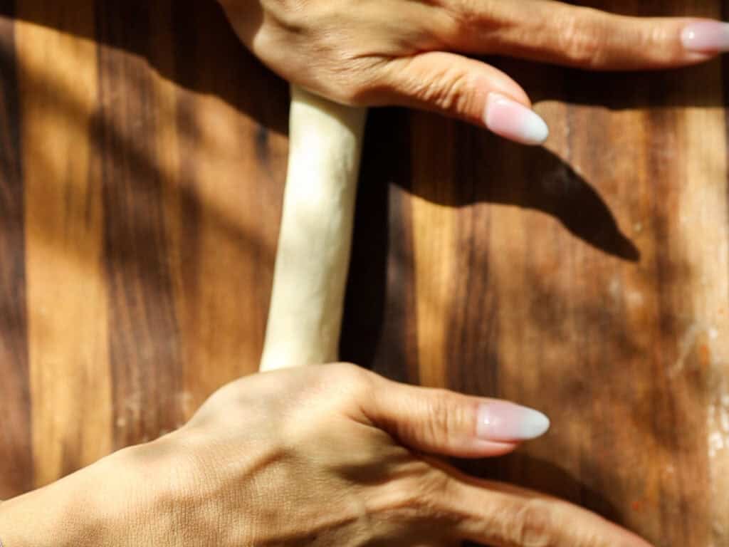Easy and Delicious Garlic Knots Recipe (Vegan) 12 Close-up of two hands with manicured nails rolling out dough on a wooden surface, with sunlight casting soft shadows.