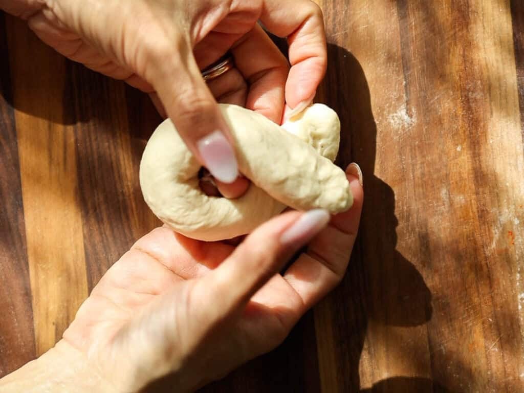 Easy and Delicious Garlic Knots Recipe (Vegan) 13 Close-up of hands shaping a piece of dough into a loop on a wooden surface, with sunlight casting soft shadows.