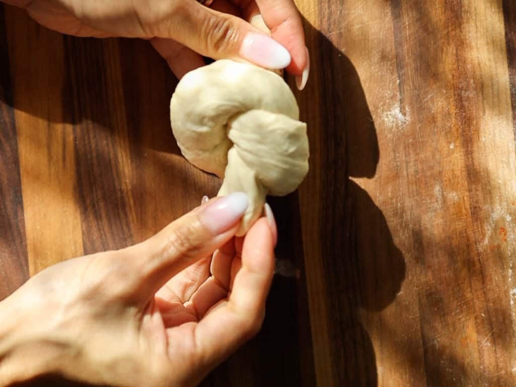 Easy and Delicious Garlic Knots Recipe (Vegan) 14 Close-up of hands shaping a piece of dough into a knot on a wooden surface, with sunlight casting shadows across the scene.