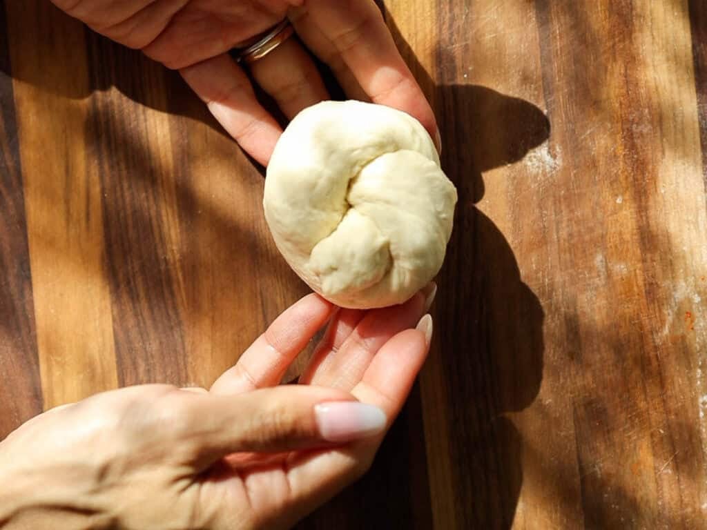 Easy and Delicious Garlic Knots Recipe (Vegan) 15 Two hands gently hold a small, round ball of dough on a wooden surface, with light and shadows visible across the scene.