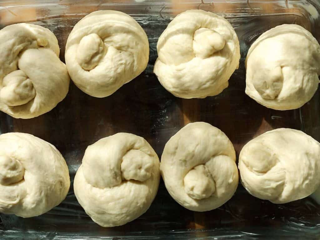 Easy and Delicious Garlic Knots Recipe (Vegan) 16 Eight unbaked dough knots are arranged in a glass baking dish, ready to be baked. The dough is smooth and lightly floured, with sunlight highlighting the texture.