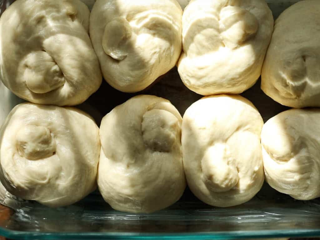 Easy and Delicious Garlic Knots Recipe (Vegan) 17 Eight balls of unbaked dough are closely packed in a glass baking dish, with sunlight highlighting their smooth, slightly glossy surfaces.