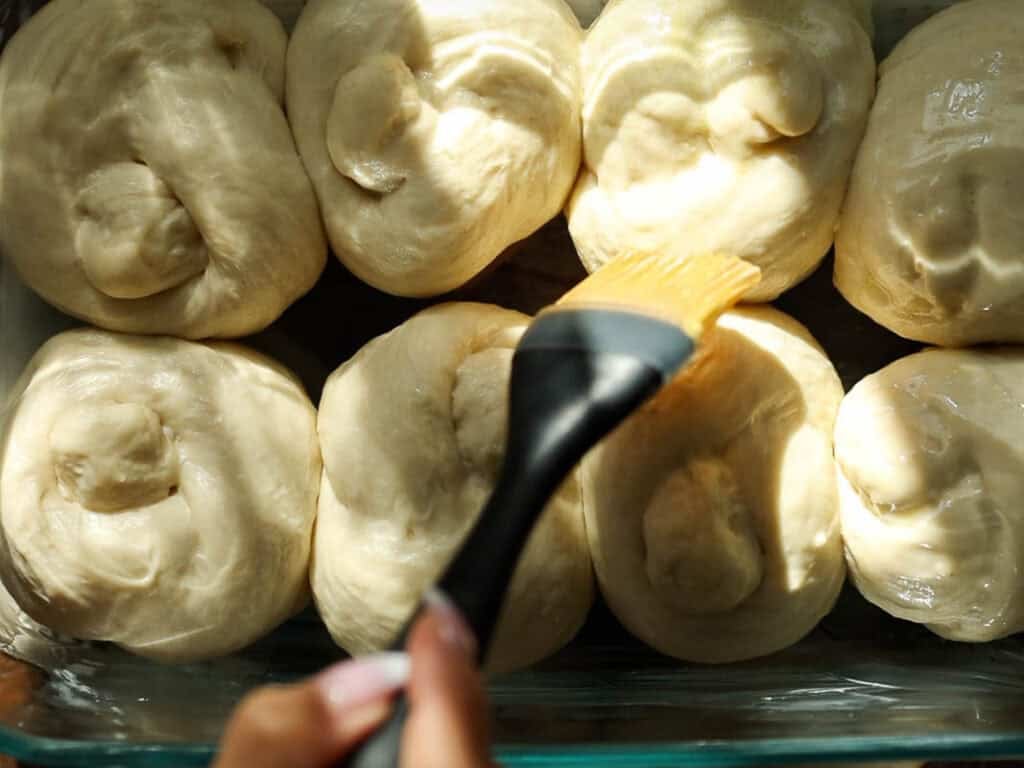 Easy and Delicious Garlic Knots Recipe (Vegan) 18 A hand uses a silicone brush to apply vegan wash to raw dough rolls arranged closely together in a baking dish, preparing them for baking.
