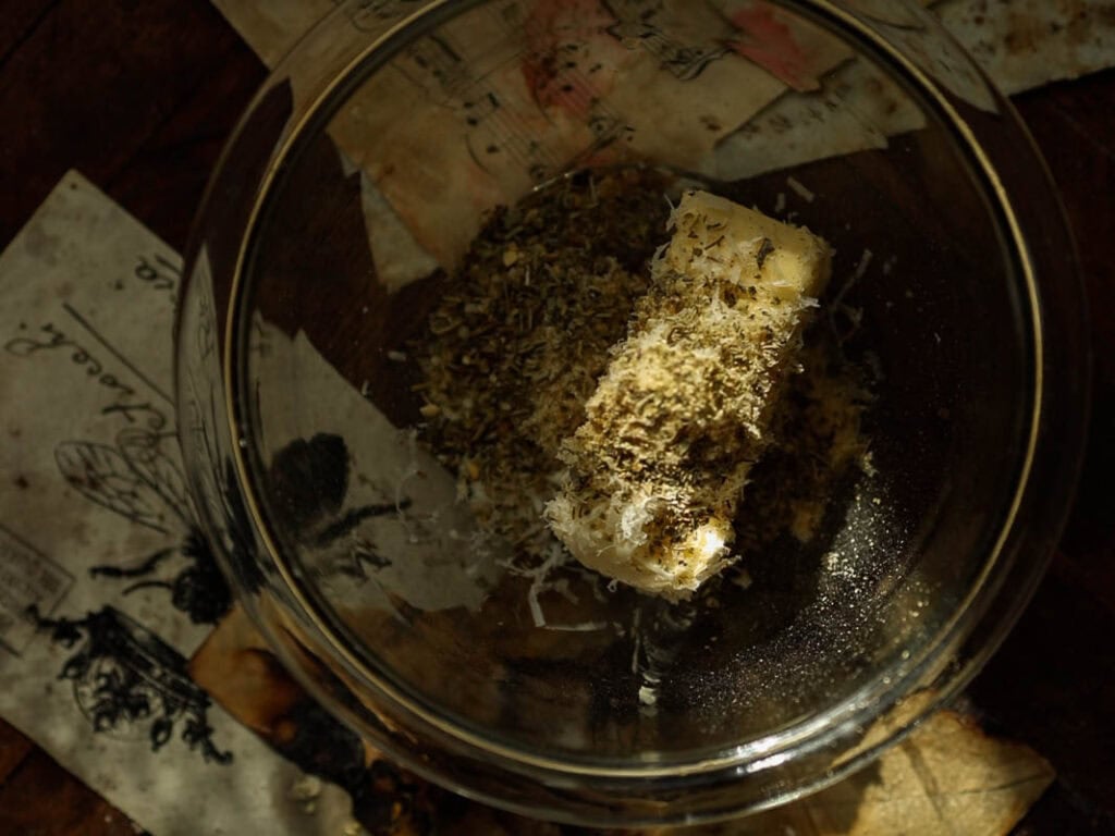 Easy and Delicious Garlic Knots Recipe (Vegan) 21 A glass bowl containing a stick of butter topped with dried herbs and shredded cheese, placed on a table with vintage-style papers featuring illustrations and musical notes.