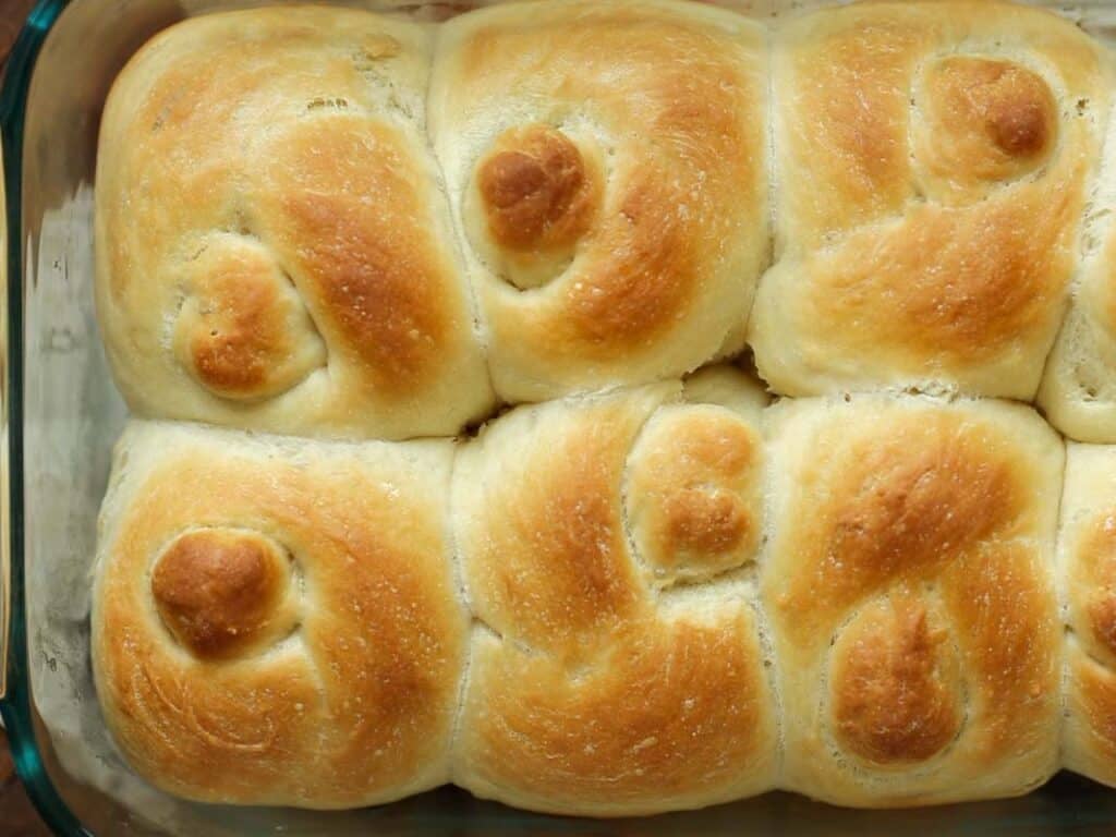 Easy and Delicious Garlic Knots Recipe (Vegan) 19 Golden brown, freshly baked dinner rolls with a swirl pattern on top, arranged closely together in a glass baking dish.