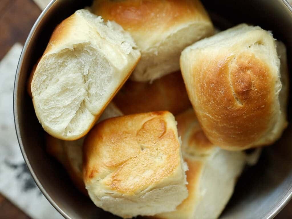 Easy and Delicious Garlic Knots Recipe (Vegan) 22 A bowl filled with several golden-brown, fluffy, homemade bread rolls, some showing soft, airy interiors where they have been pulled apart.
