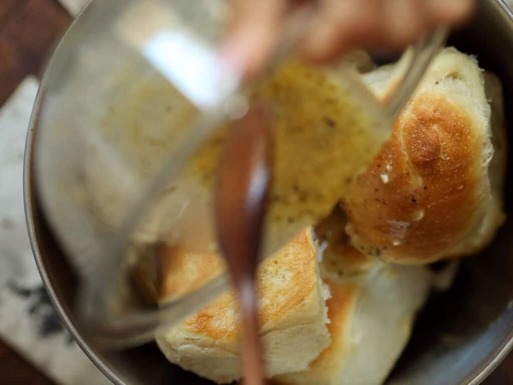Easy and Delicious Garlic Knots Recipe (Vegan) 23 A close-up of golden brown dinner rolls in a bowl, with a hand pouring melted butter over them using a spoon.