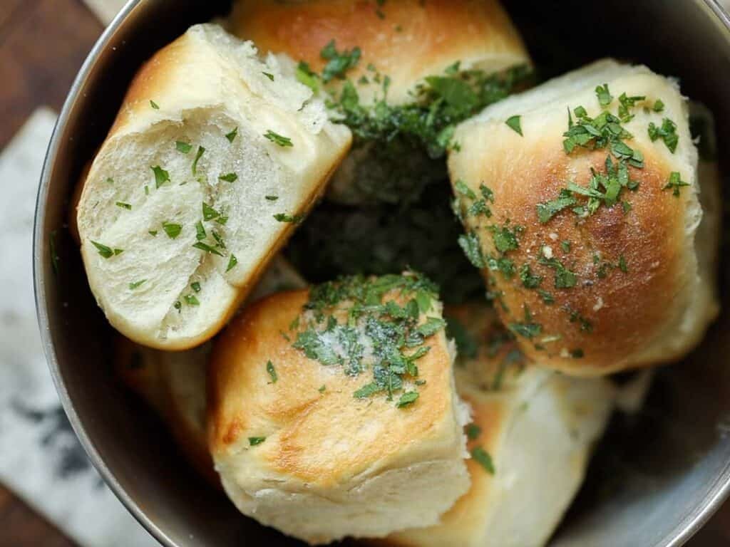 Easy and Delicious Garlic Knots Recipe (Vegan) 24 A close-up of soft, golden brown dinner rolls in a bowl, topped with chopped fresh parsley and a sprinkle of coarse salt.