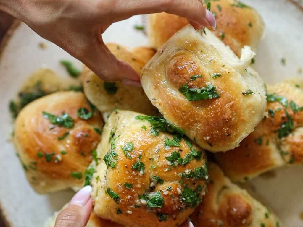 Easy and Delicious Garlic Knots Recipe (Vegan) 3 A hand reaches for a golden, fluffy garlic knots recipe topped with chopped fresh herbs, surrounded by several similar rolls on a plate.
