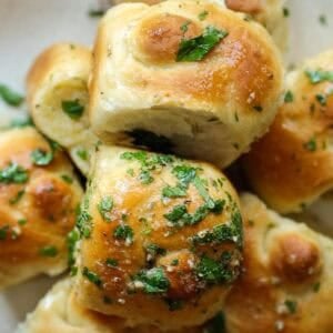 Easy and Delicious Garlic Knots Recipe (Vegan) 29 A close-up of golden, fluffy garlic knots recipe topped with chopped parsley and sprinkled with grated cheese, showing a soft, airy interior and a shiny, buttery surface.