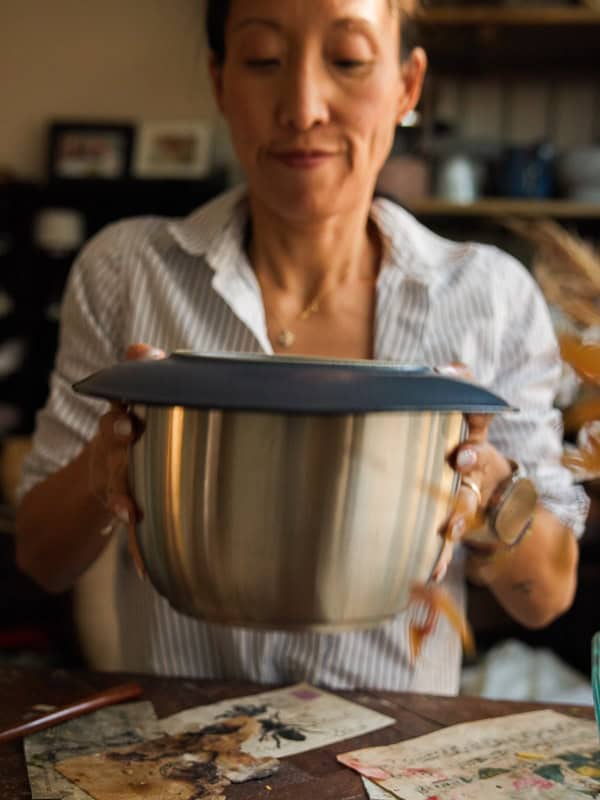 Easy and Delicious Garlic Knots Recipe (Vegan) 25 A woman in a striped shirt holds a large metal mixing bowl covered with a dark lid, standing at a table with papers and dried leaves scattered on it. She appears to be focused on her task.