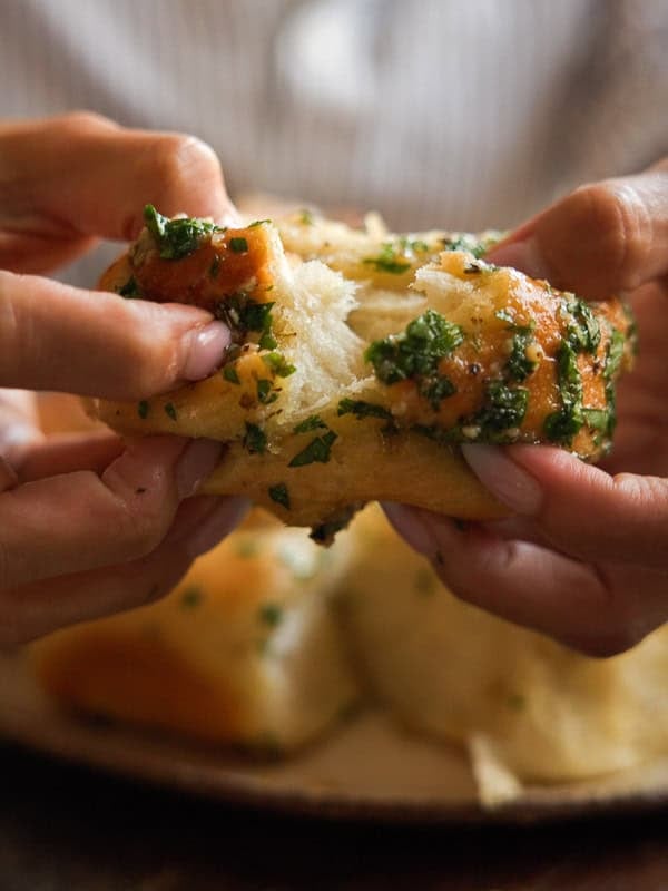 Easy and Delicious Garlic Knots Recipe (Vegan) 2 Close-up of hands pulling apart a piece of soft, fluffy garlic bread coated with chopped herbs and butter, with more bread in the blurred background.