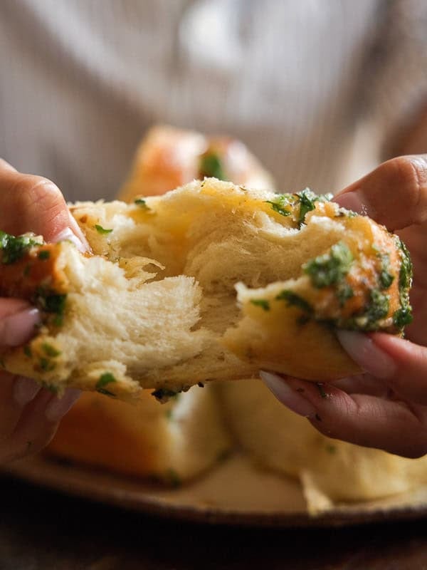 Easy and Delicious Garlic Knots Recipe (Vegan) 27 A person pulls apart a piece of fluffy bread topped with chopped herbs, showing the soft, airy texture inside.