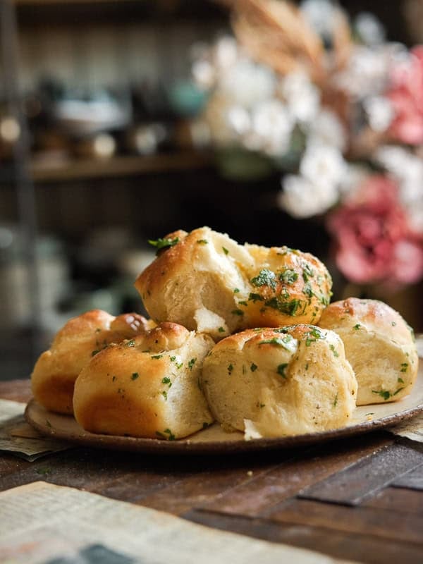Easy and Delicious Garlic Knots Recipe (Vegan) 26 A plate of freshly baked garlic knots recipe sits on a wooden table, with a cozy, blurred background featuring flowers and kitchenware.