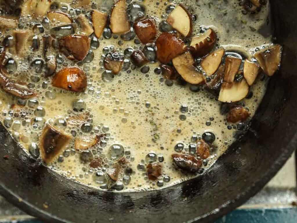 15-minute Quick and Easy Vegan Mushroom Gravy Recipe. 3 Chopped mushrooms cooking in a skillet with bubbling liquid, possibly butter or oil, creating a foamy texture around the pieces.