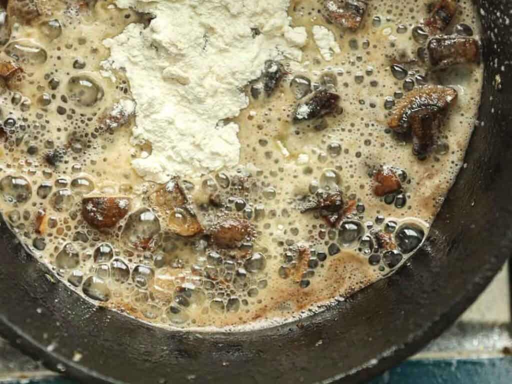 15-minute Quick and Easy Vegan Mushroom Gravy Recipe. 4 A close-up of a skillet with bubbling liquid, sautéed mushrooms, and a mound of flour in the center, starting to mix together for a recipe.