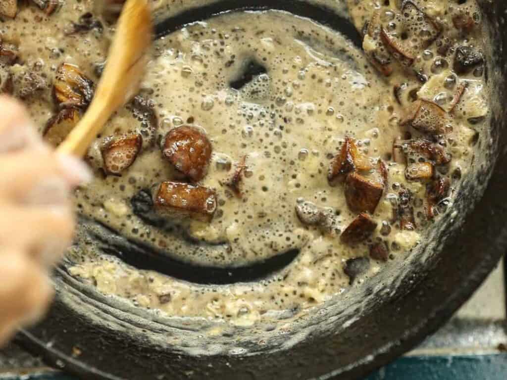 15-minute Quick and Easy Vegan Mushroom Gravy Recipe. 5 A close-up of a skillet with bubbling sauce and chunks of browned food being stirred with a wooden spoon, creating a smiley face pattern in the mixture.