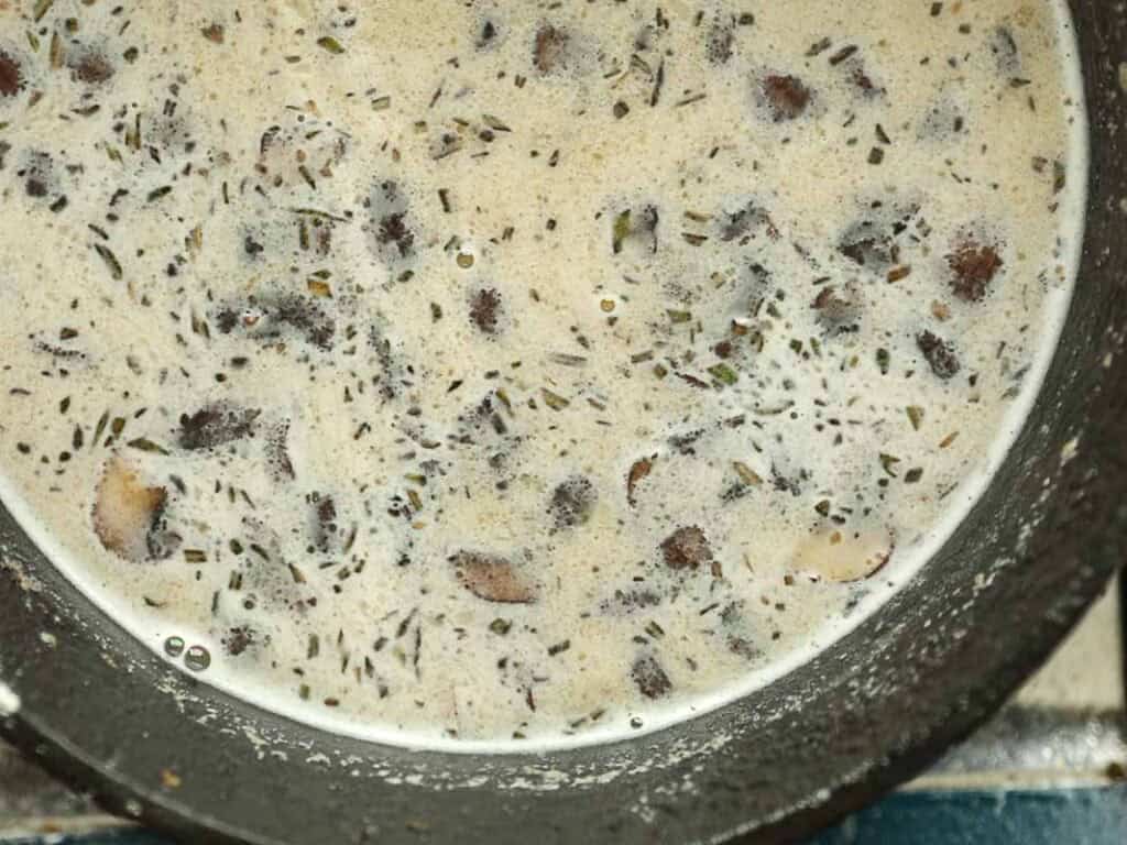 15-minute Quick and Easy Vegan Mushroom Gravy Recipe. 6 Close-up of a creamy vegan mushroom liquid with visible chunks of mushrooms and herbs, served in a black bowl. The soup has a speckled appearance from the herbs and mushroom pieces.