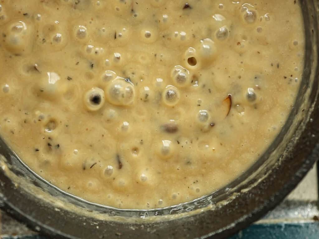 15-minute Quick and Easy Vegan Mushroom Gravy Recipe. 7 A close-up view of creamy, bubbling creamy vegan mushroom gravy with visible grains and specks of spices in a bowl.