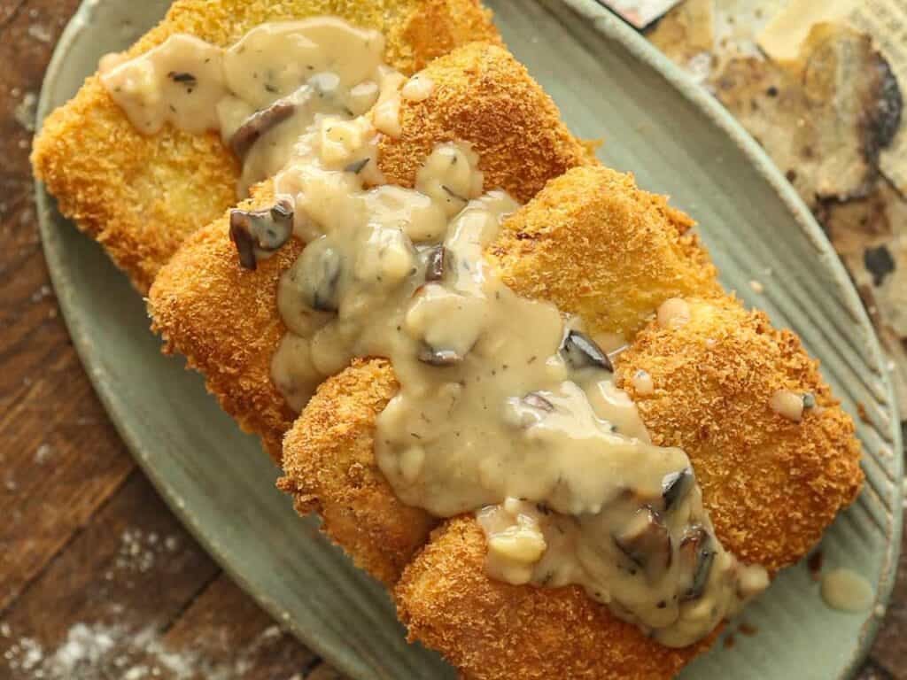 15-minute Quick and Easy Vegan Mushroom Gravy Recipe. 2 A breaded, golden-brown fried cutlet topped with creamy vegan mushroom sauce is served on an oval plate, set on a rustic wooden surface.