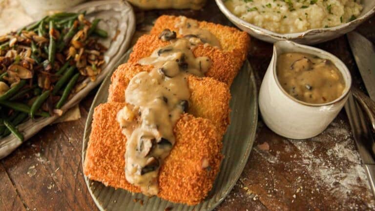 Recipes 13 Three breaded and fried cutlets topped with creamy vegan mushroom gravy on an oval plate, with a side of green beans, mashed potatoes, and extra gravy in a small pitcher on a rustic wooden table.