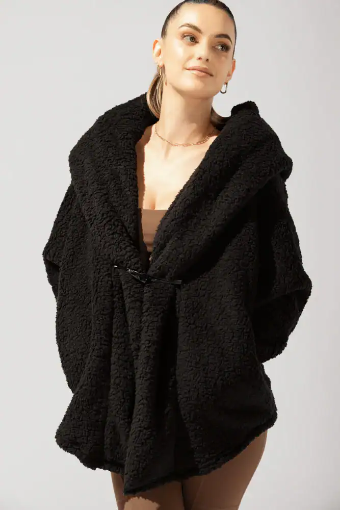 The Korean Vegan's 2025 VEGAN Holiday Gift GUIDE. 42 A woman with pulled-back hair wears a cozy, oversized black sherpa jacket—a perfect Korean Vegan Holiday Gift for 2025—with a hood, brown top and pants, and hoop earrings. She smiles slightly, looking to the side against a plain background.