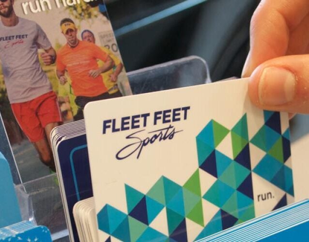 The Korean Vegan's 2025 VEGAN Holiday Gift GUIDE. 38 A hand selects a Fleet Feet Sports gift card from a display rack filled with colorful options, perfect for any 2025 Vegan Holiday Gift Guide or thoughtful present for your favorite Korean Vegan runner.
