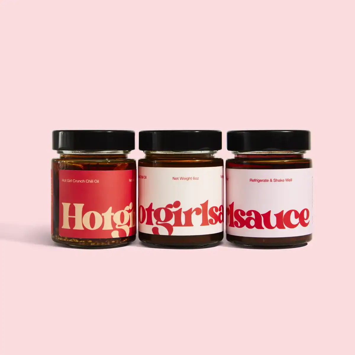 The Korean Vegan's 2025 VEGAN Holiday Gift GUIDE. 45 Three glass jars with black lids containing hot sauce, arranged side by side on a light pink background. Perfect as a Vegan Holiday Gift for 2025, their labels display bold red and white text reading Hot Girl Sauce and Hot Girl Crunch Chili Oil.