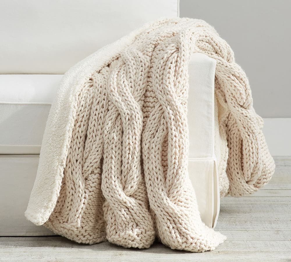 The Korean Vegan's 2025 VEGAN Holiday Gift GUIDE. 40 A chunky, cream-colored cable knit blanket is draped over the arm of a white upholstered chair with light wood flooring below—perfect for your Vegan Holiday Gift Guide 2025 or cozy Korean Vegan-inspired moments.