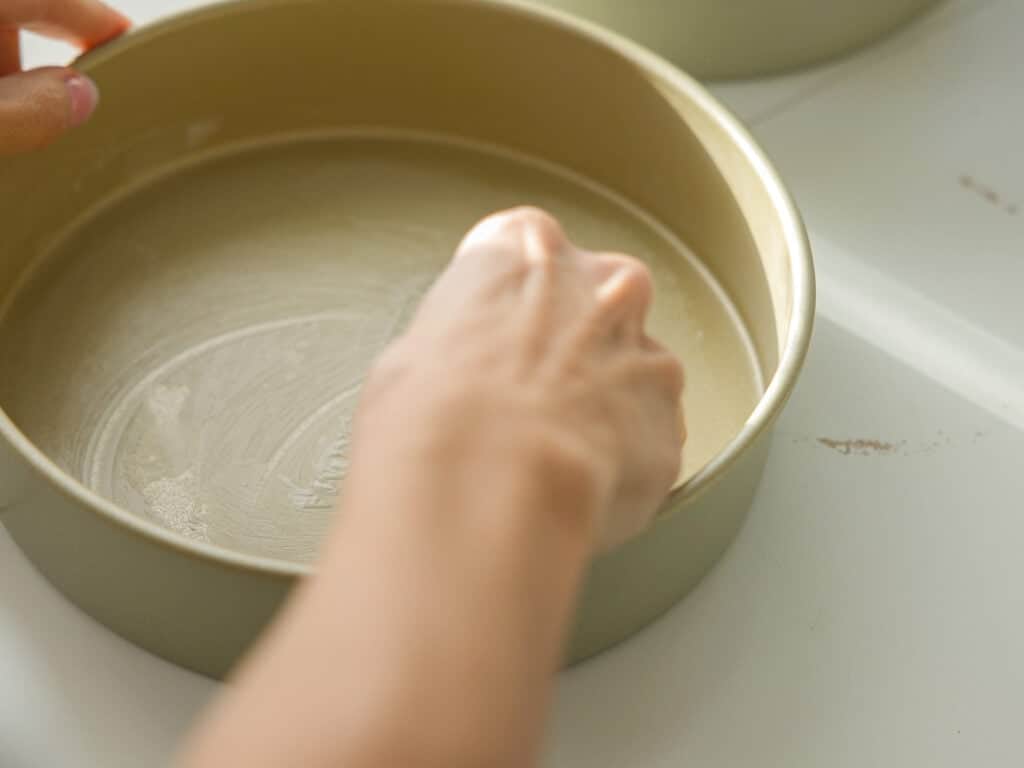 The BEST Vegan Chocolate Cake Recipe On The Internet. 6 A close-up of a persons hand greasing a round cake pan with butter or oil, preparing it for baking. The pan is on a light-colored surface.