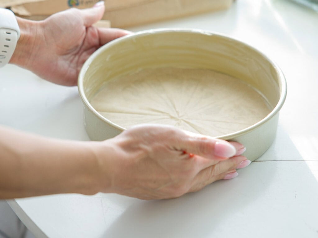 The BEST Vegan Chocolate Cake Recipe On The Internet. 7 A person holding a round baking pan lined with parchment paper, preparing it for baking on a white countertop.