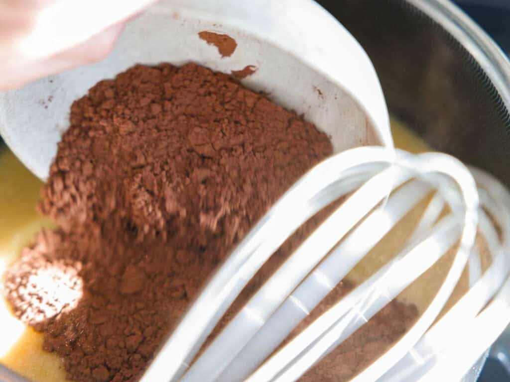 The BEST Vegan Chocolate Cake Recipe On The Internet. 9 A close-up of cocoa powder being poured from a bowl into a mixing bowl with a whisk, ready to be combined with other ingredients. Bright lighting highlights the rich brown and yellow colors.