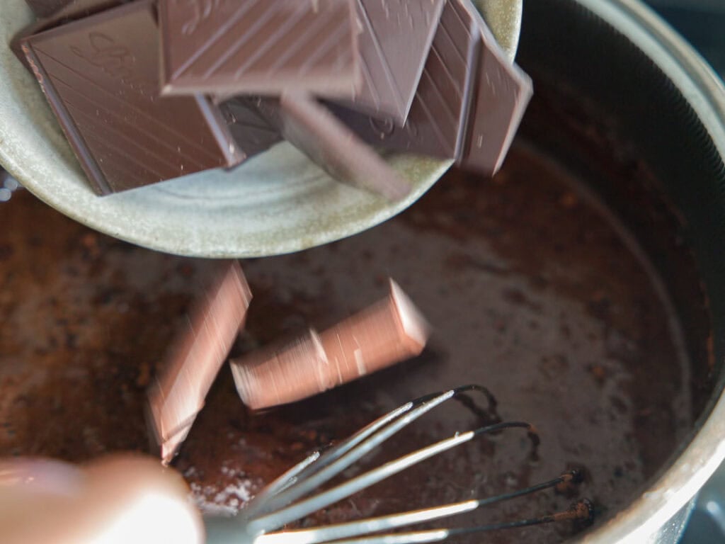 The BEST Vegan Chocolate Cake Recipe On The Internet. 10 A close-up of chocolate pieces being poured from a bowl into a saucepan of melted chocolate mixture, with a whisk stirring the mixture.
