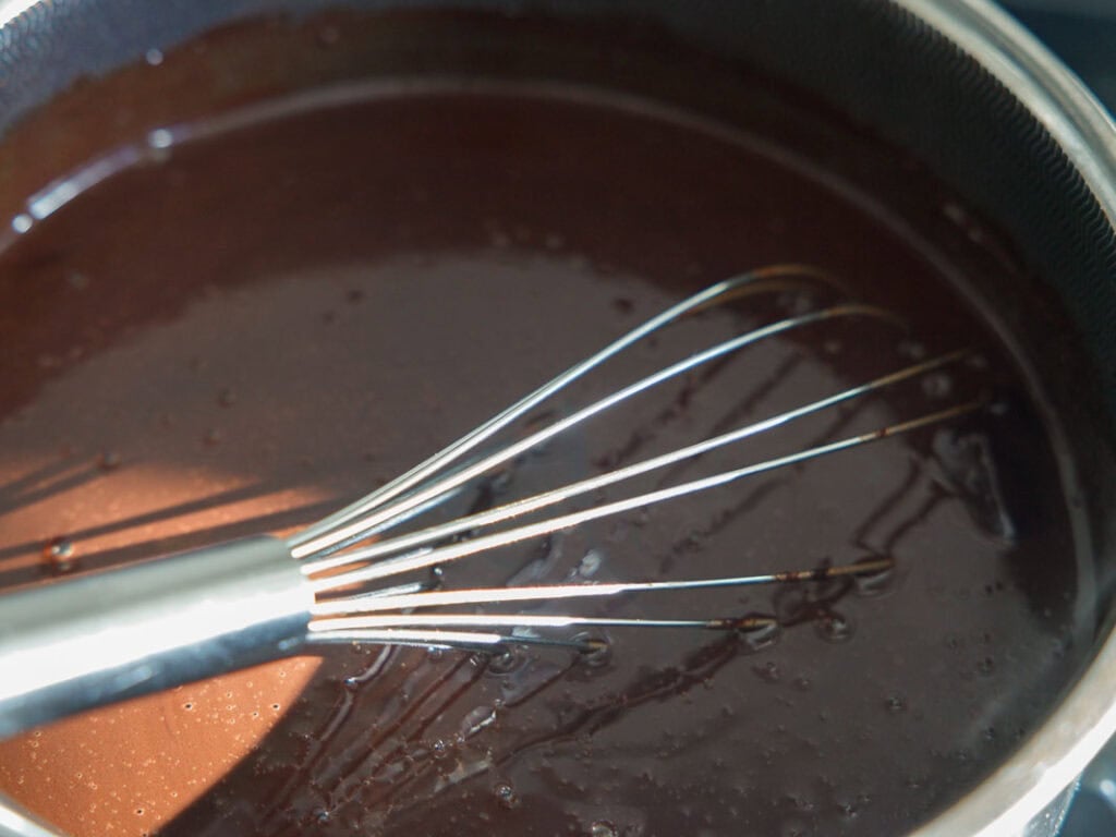 The BEST Vegan Chocolate Cake Recipe On The Internet. 12 A metal whisk is mixing glossy, melted chocolate in a dark, round pan, creating a smooth mixture with a few small bubbles visible on the surface.