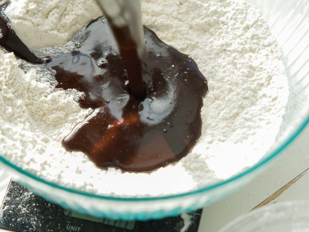 The BEST Vegan Chocolate Cake Recipe On The Internet. 13 Chocolate batter is being poured into a bowl of dry flour mixture, likely as part of a baking process. The bowl is clear and sits on a kitchen scale.