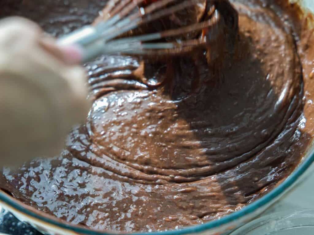 The BEST Vegan Chocolate Cake Recipe On The Internet. 15 A close-up of a hand using a whisk to mix thick, glossy chocolate batter in a clear glass bowl.