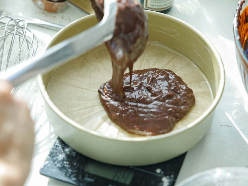 The BEST Vegan Chocolate Cake Recipe On The Internet. 16 A close-up of chocolate cake batter being spread onto parchment paper in a round baking pan, which sits on a digital kitchen scale. A whisk and bowl are visible in the background.