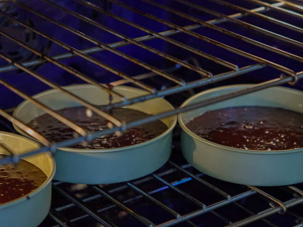 The BEST Vegan Chocolate Cake Recipe On The Internet. 17 Three round cake pans filled with chocolate batter are placed on an oven rack, ready to be baked. The oven rack and the open oven interior are visible.