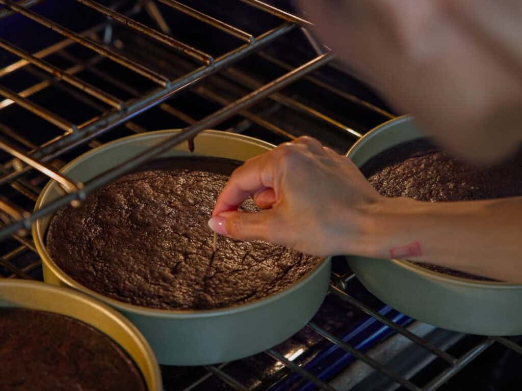 The BEST Vegan Chocolate Cake Recipe On The Internet. 18 A person tests the doneness of a chocolate cake in a round pan with a toothpick inside an oven. Two additional cakes in similar pans are also visible on the oven rack.